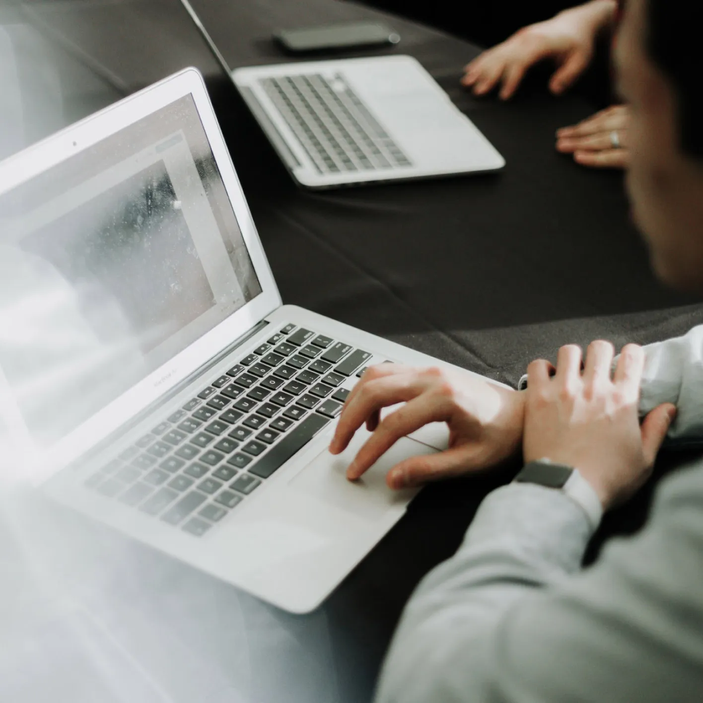 Person using a laptop with another closed laptop and two hands resting on a black table in the background.